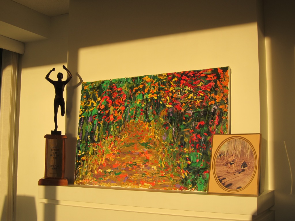 The photo shows a large abstract painting with bright forest leaves. On the left is a large trophy showing a woman runner with her hands raised in triumph. On the right is a framed photo showing a solitary female runner against bare November trees, with one spectator nearby and others in the distance.